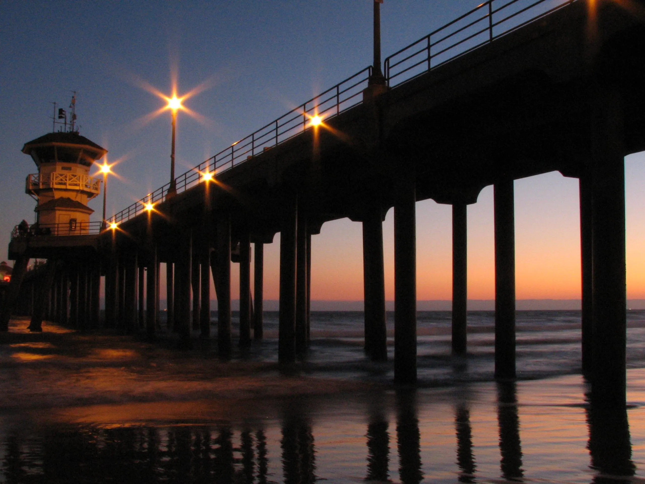 Huntington Beach Pier at twilight with lit lampposts reflecting on wet sand.