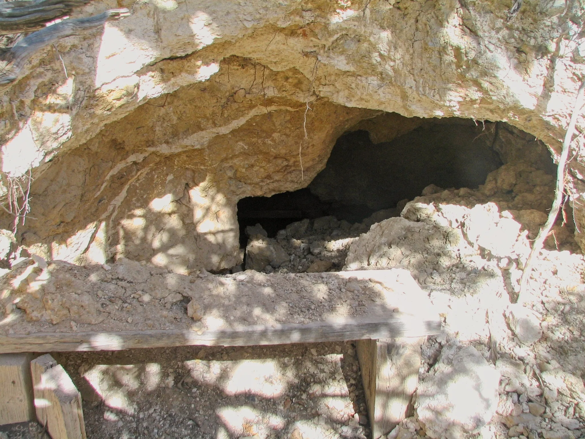 Mine portal under a rocky overhang with a dirt-covered wooden bench in front.