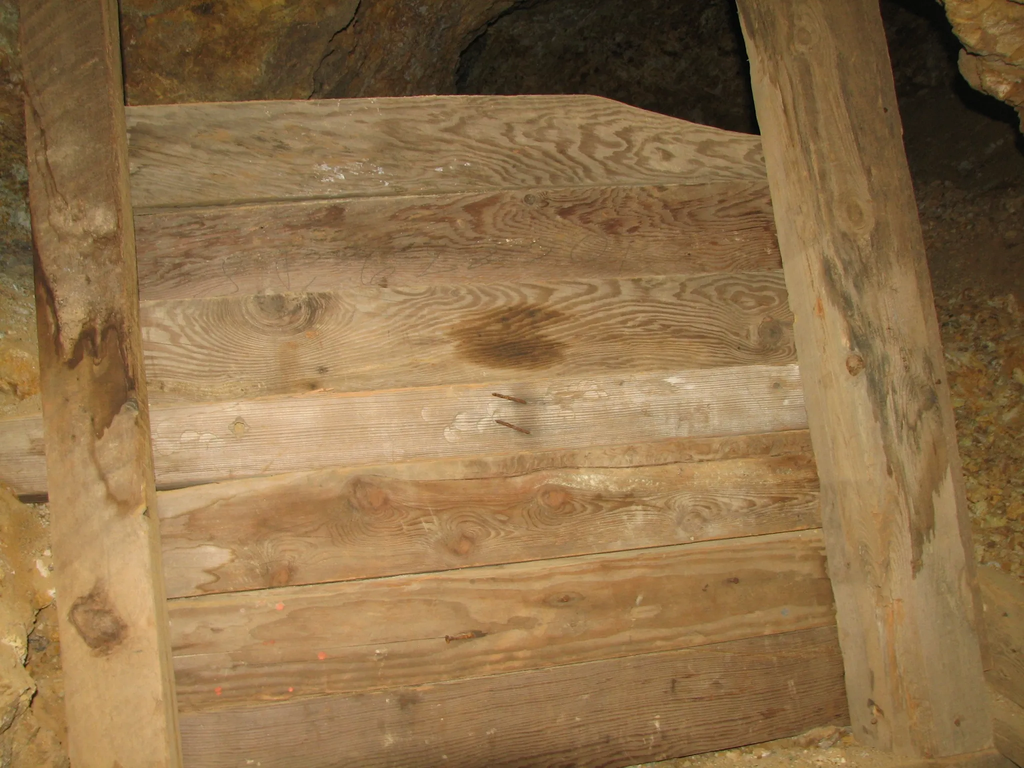 Horizontal wooden planks blocking a mine passage with rusted nails and rock walls.