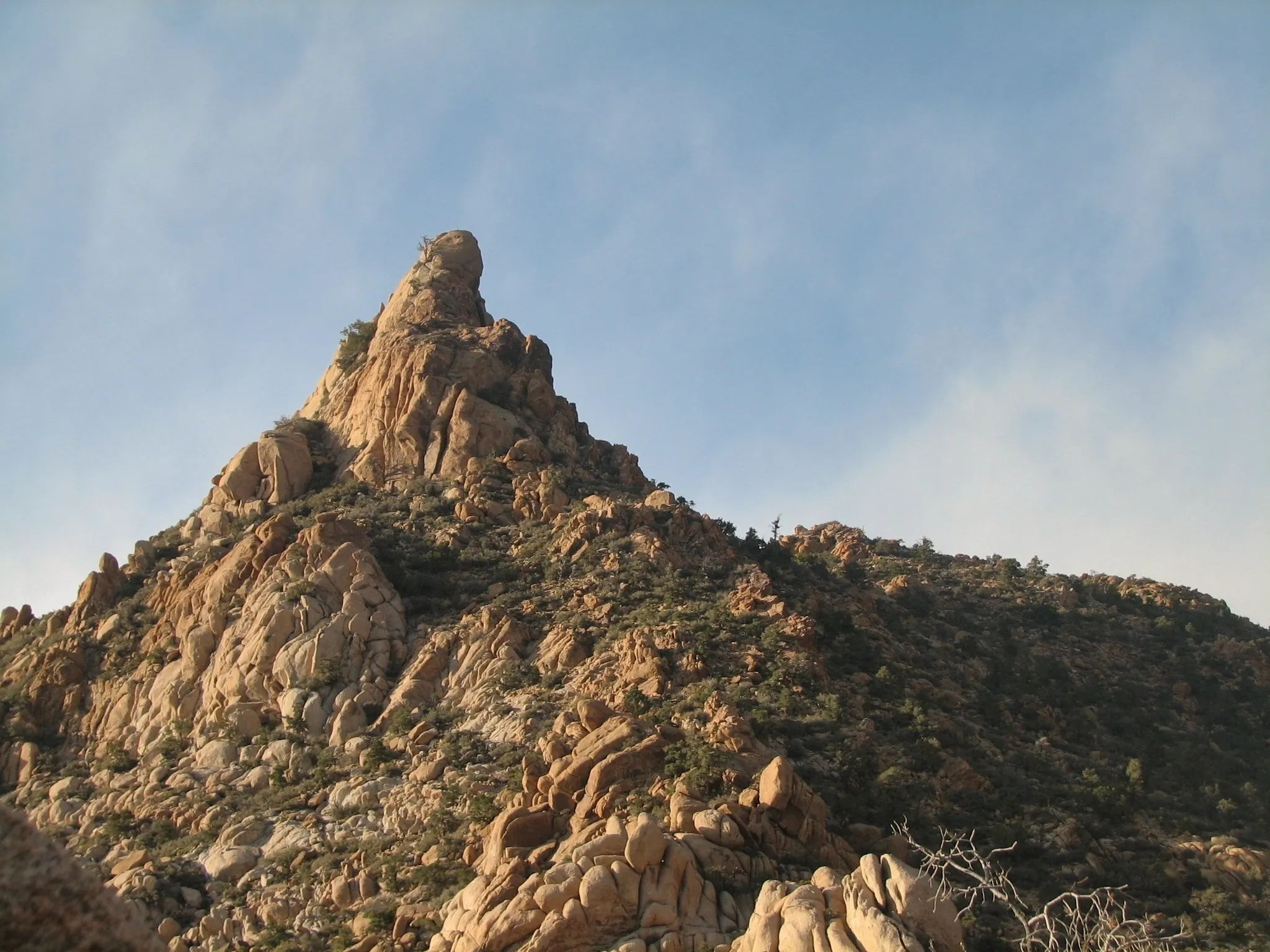 Jagged tan granite peak with sparse shrubs under a pale blue desert sky.