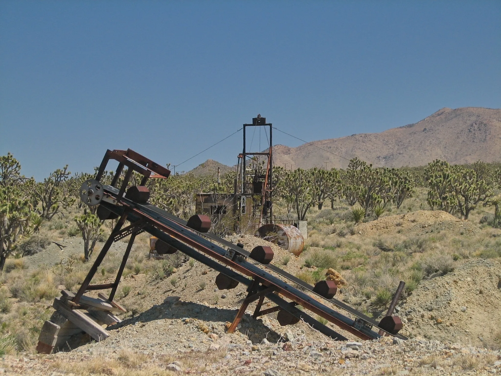 Rusted inclined conveyor belt, mine headframe, and tank on sandy tailings.