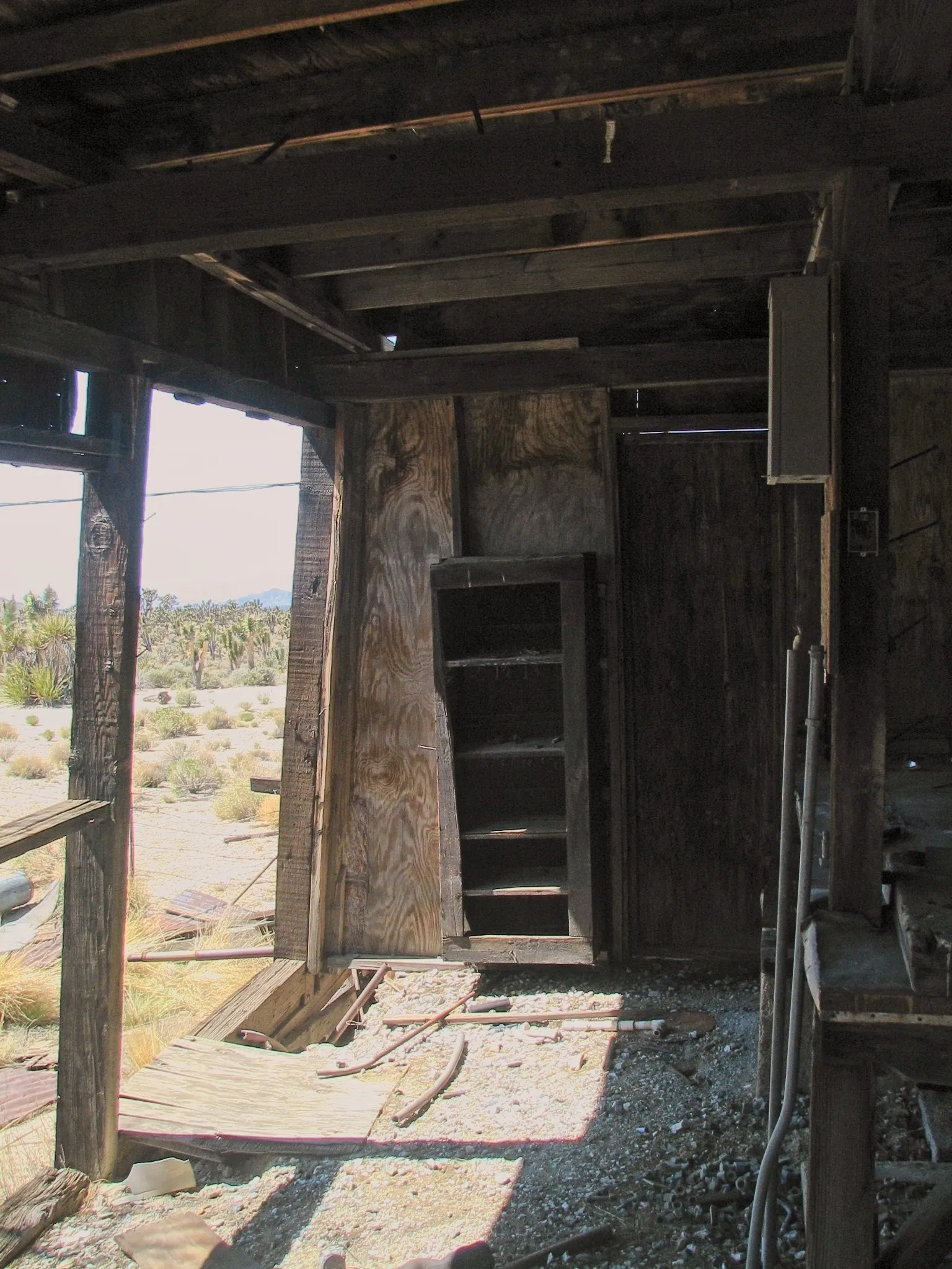 Interior of abandoned wooden shack with bookshelf and desert view through opening.
