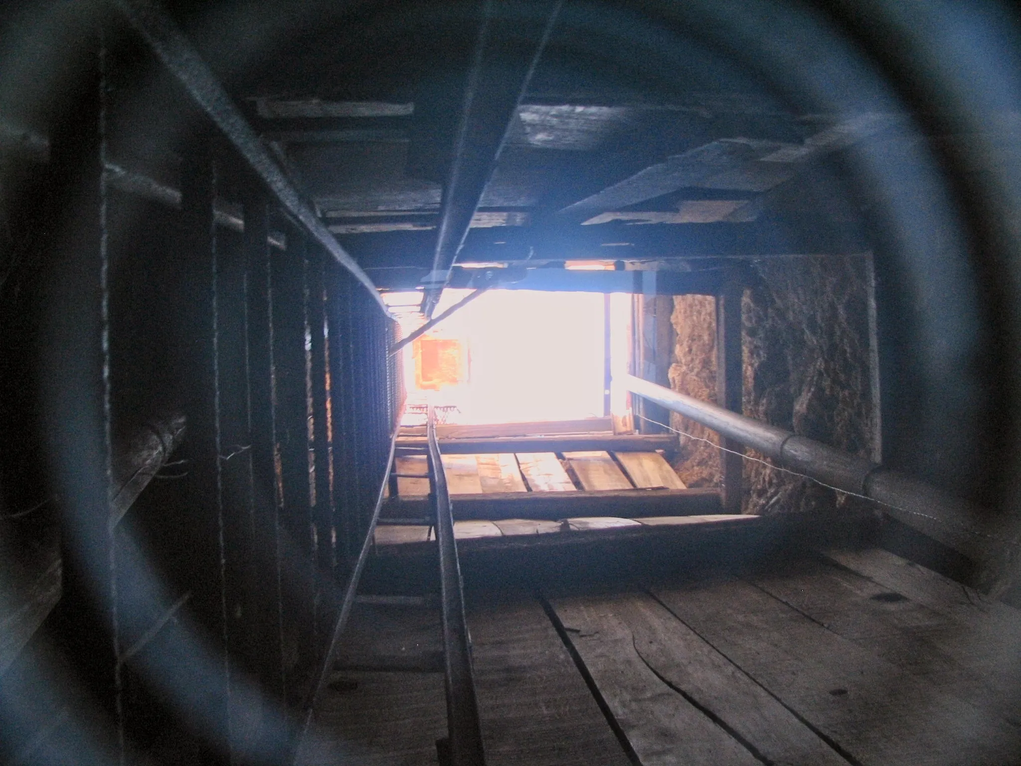 Looking up a mine shaft toward daylight with ladder and pipes.