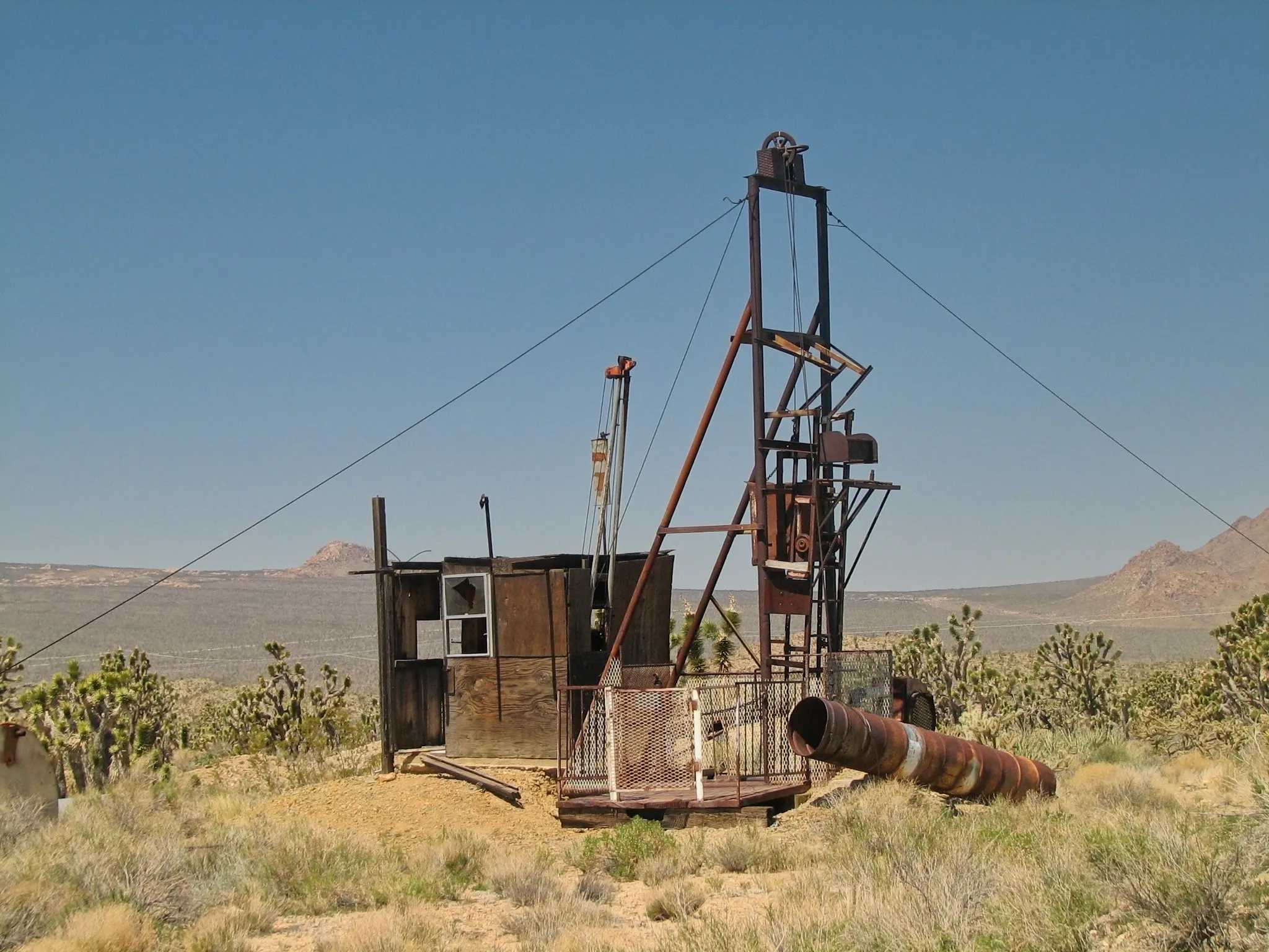 Rusted mine headframe and wooden shack with Joshua trees behind.