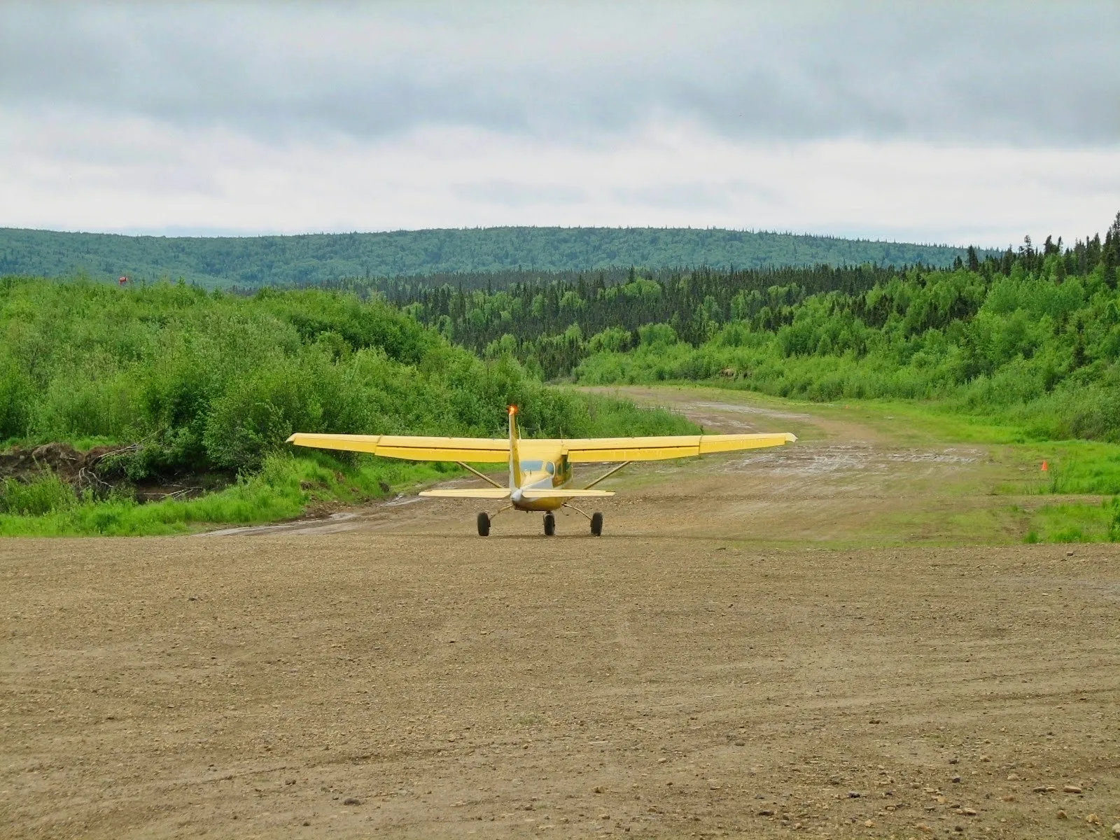 Yellow Cessna 206 on a gravel runway in Kako, Alaska near Russian Mission, surrounded by forested hills.