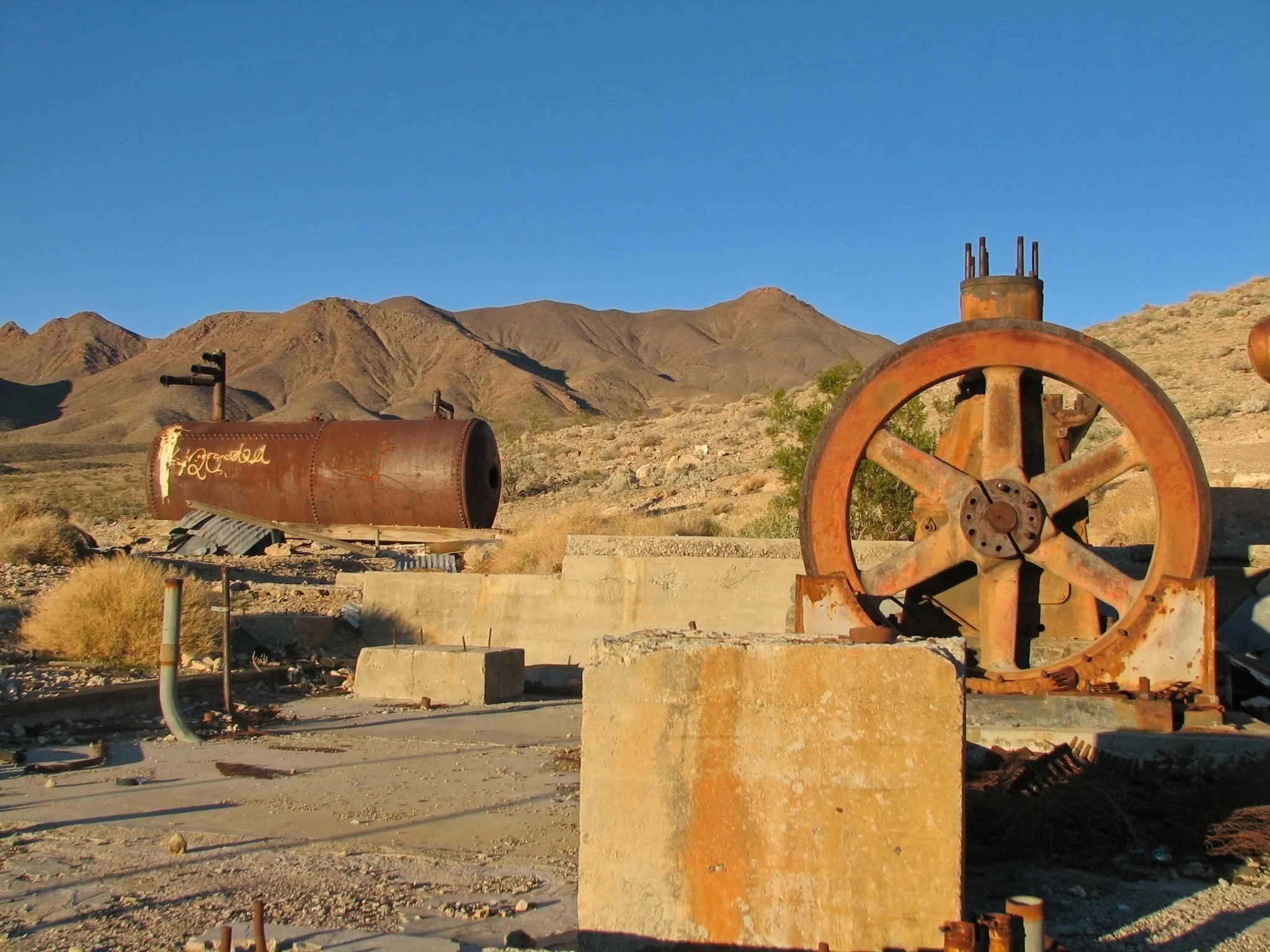 Rusted boiler and flywheel on concrete slabs with desert mountains behind.