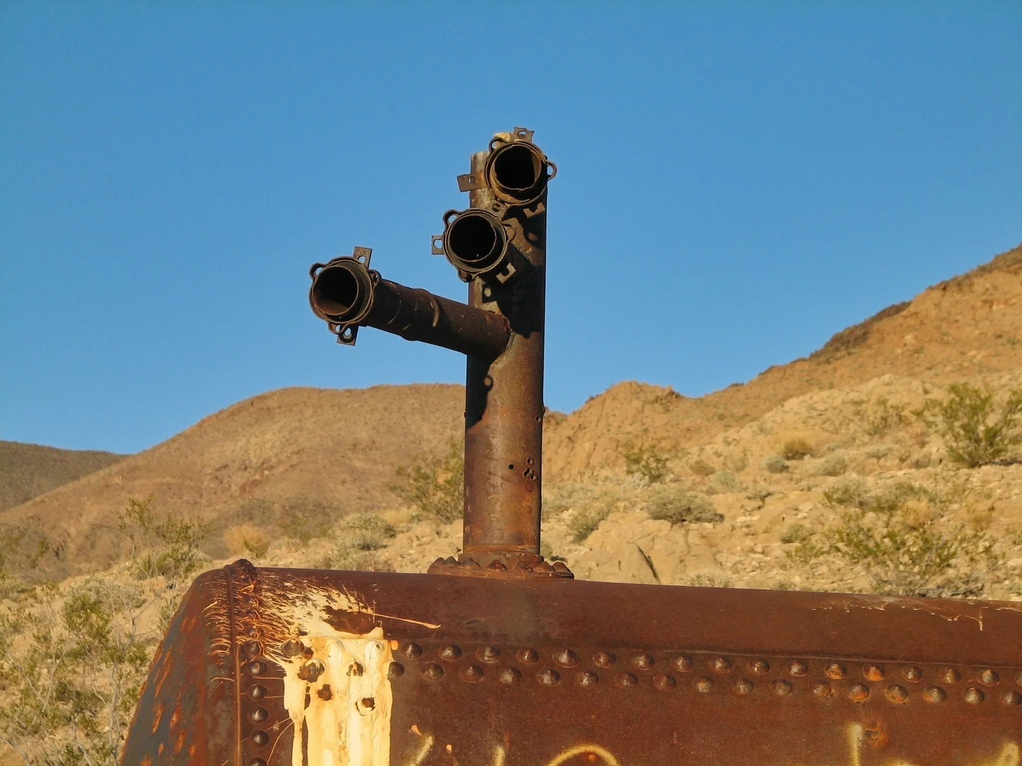 Rusted pipe nozzles on riveted tank with desert hills behind.