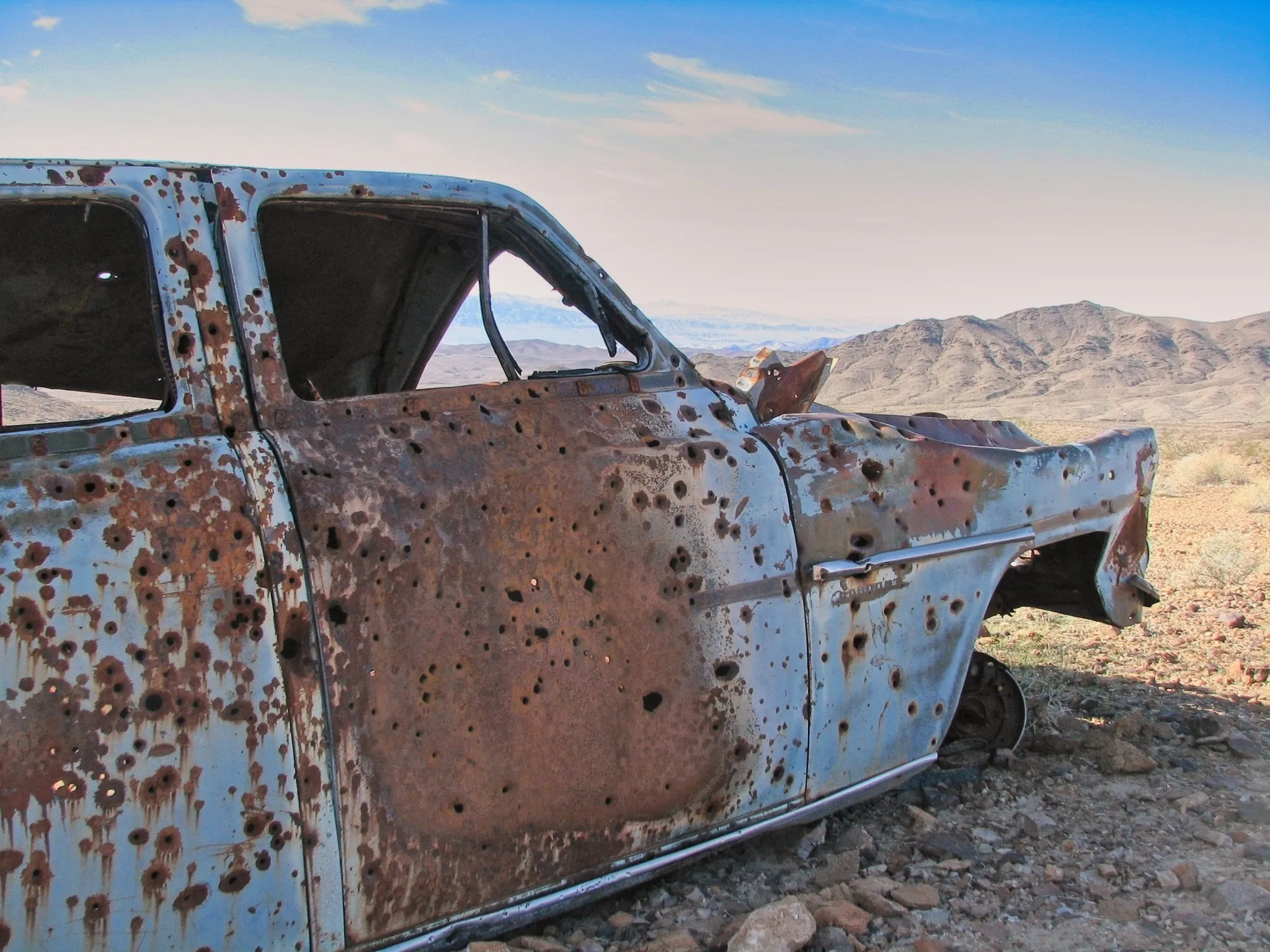Side view of bullet-riddled abandoned sedan in Mojave Desert.