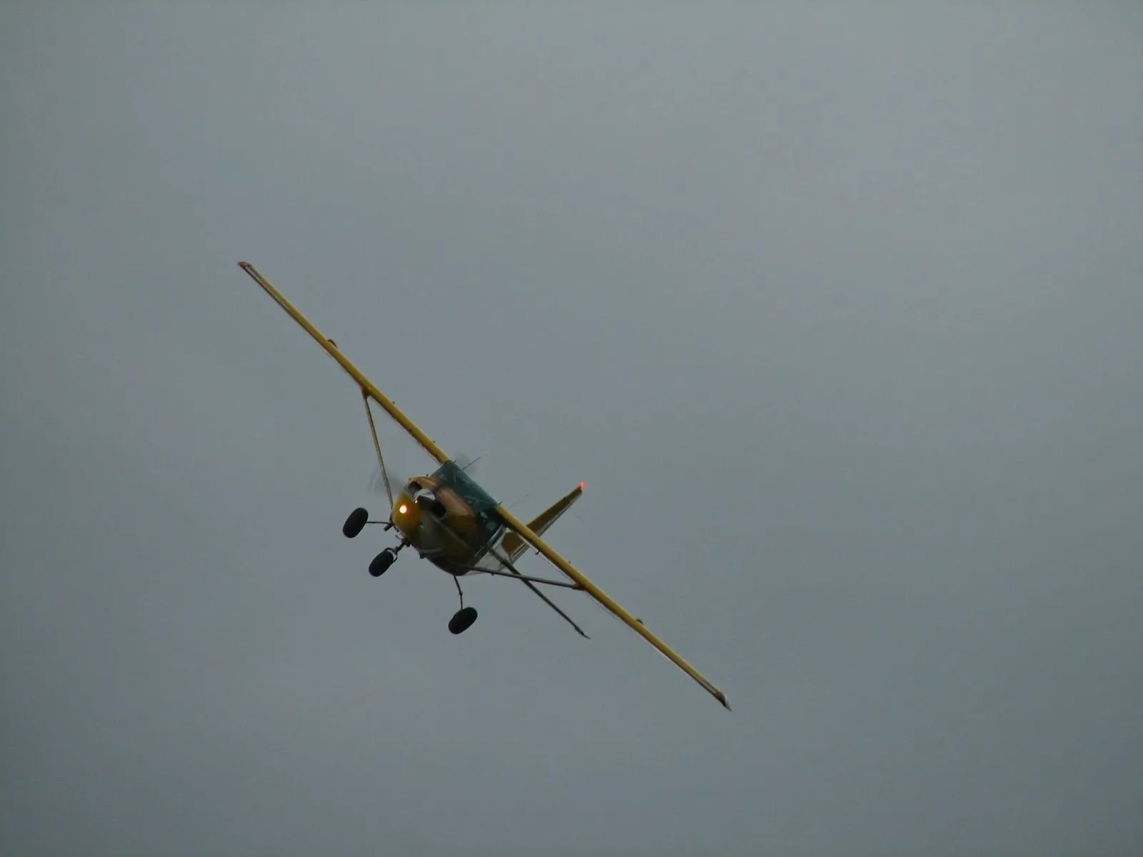 Yellow Cessna 206 banking in flight over bush Alaska near Kako, with landing light on against a gray sky.