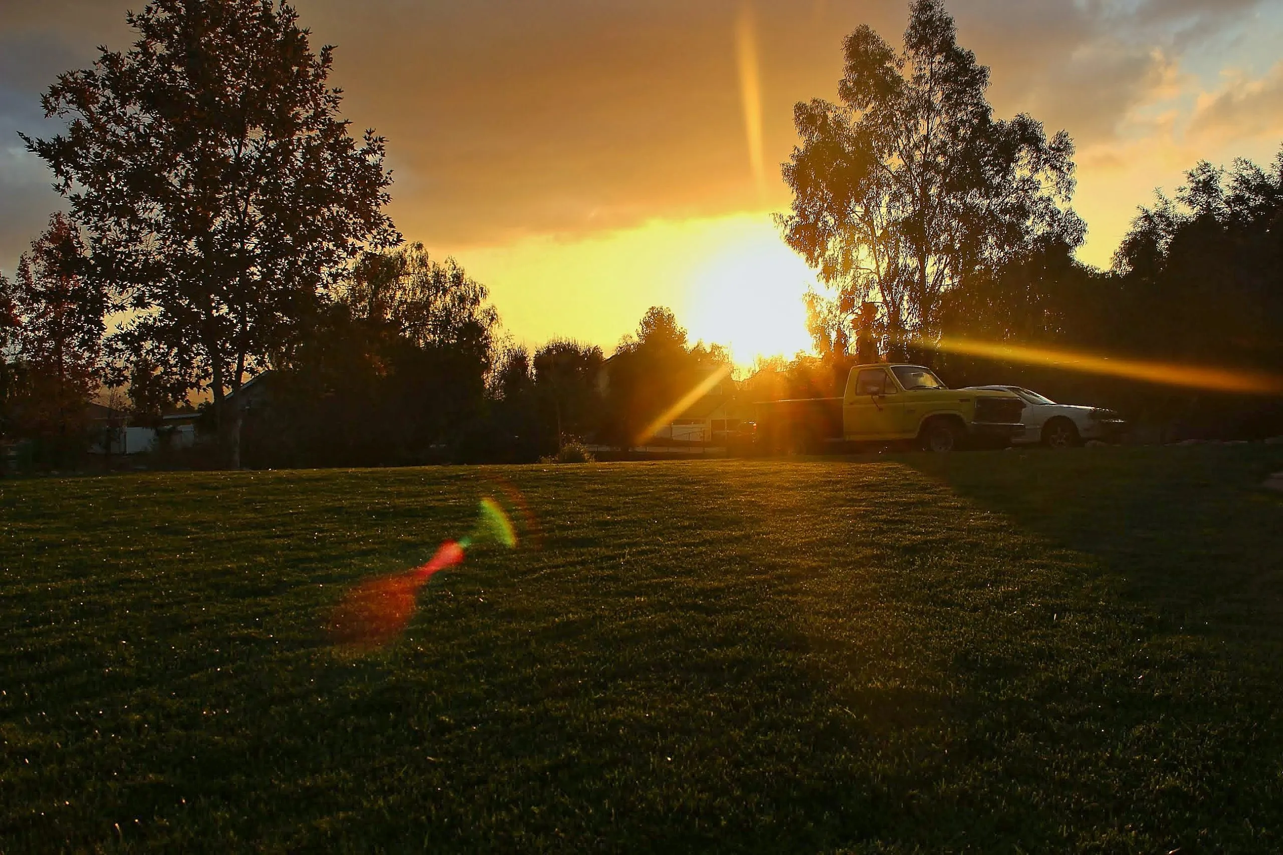 Yellow pickup truck and white sedan on a grassy field at sunset with lens flare, Woodcrest, CA.