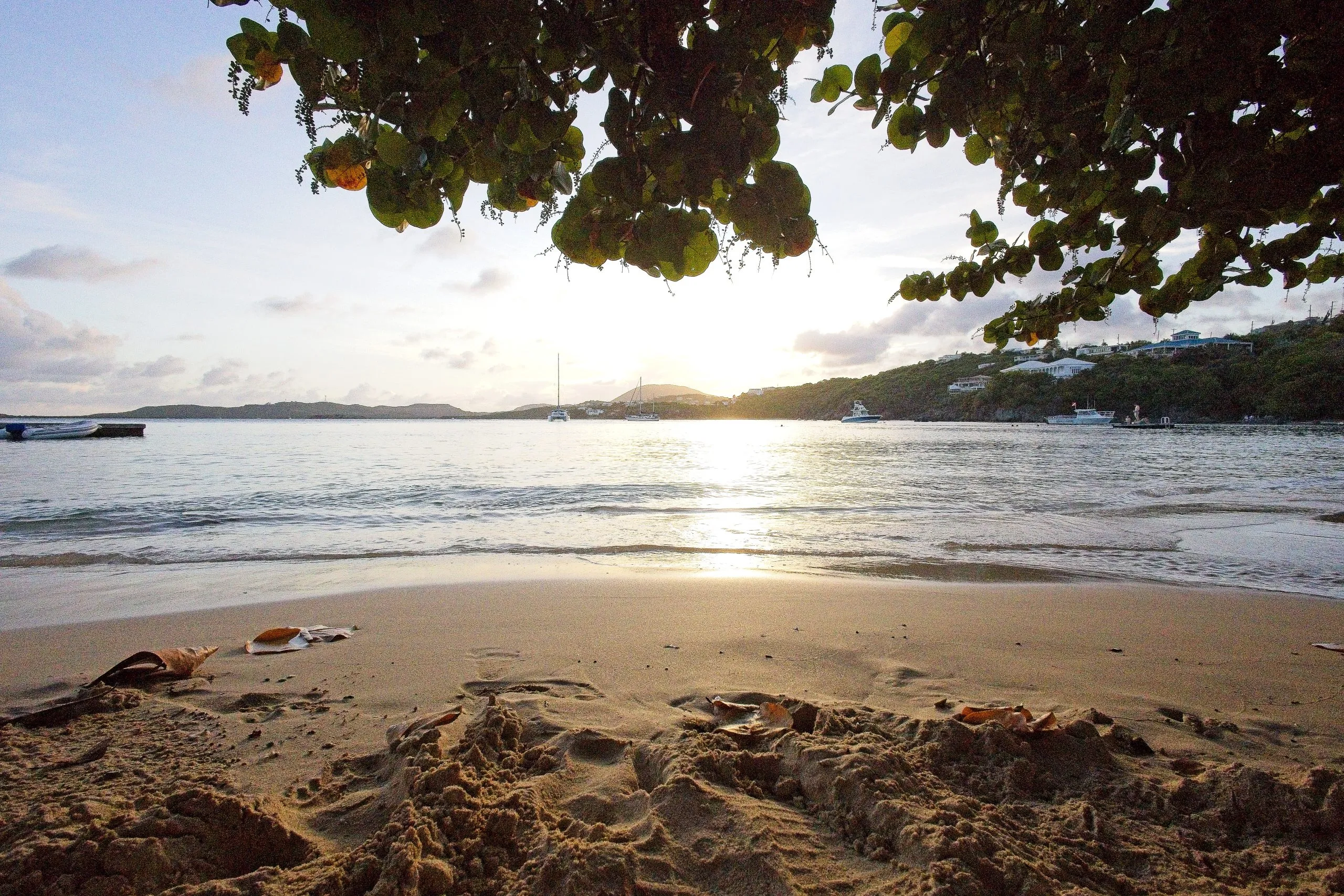 Sandy beach at Secret Harbour at sunset with anchored boats and hillside houses under overhanging tree branches, St Thomas, USVI.