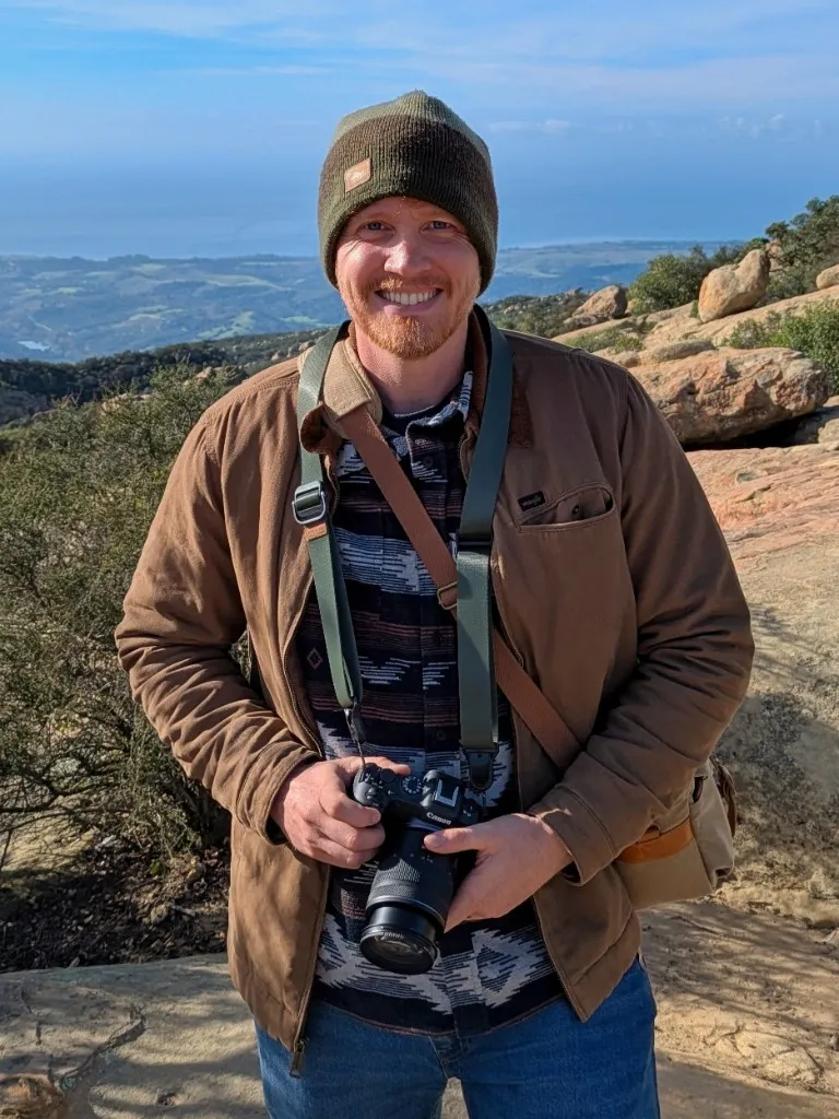 Andrew Stevens holding a Canon camera on a rocky hillside overlooking the coast
